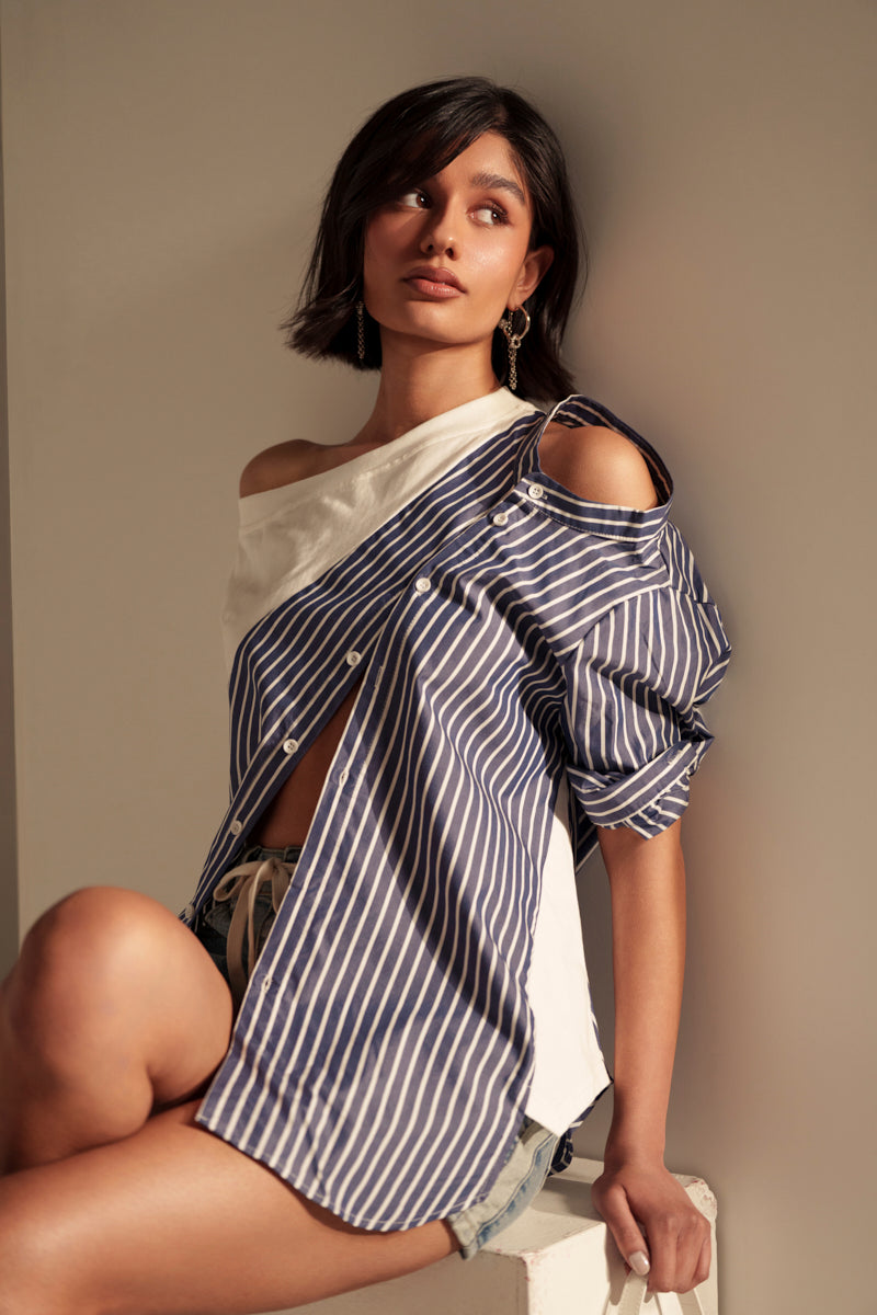 A woman with short dark hair sits on a bench, wearing the MIXED UP TSHIRT BLOUSE over a white top and shorts. She looks slightly upward with a thoughtful expression, softly lit.