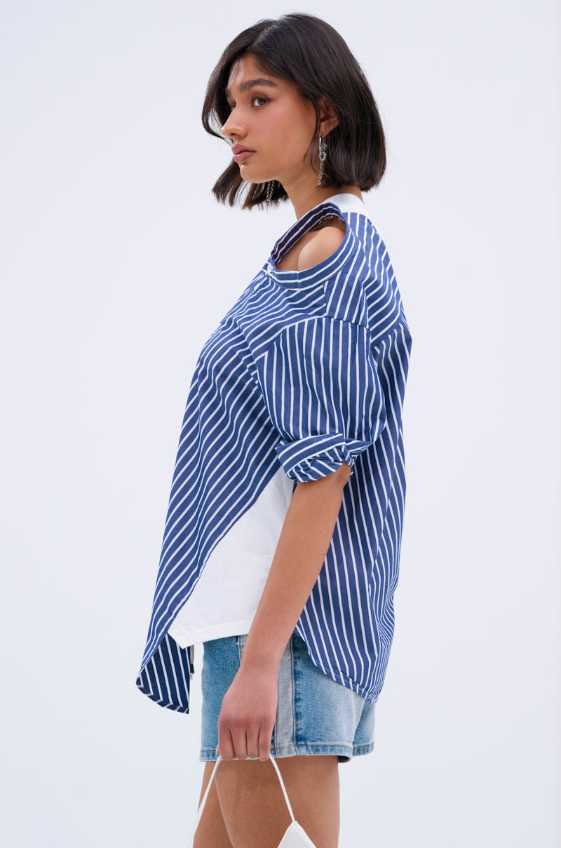 A woman with short dark hair models the MIXED UP TSHIRT BLOUSE, a blue and white striped top with shoulder cut-outs over a white layer, paired with blue denim shorts, standing sideways against a plain light background.