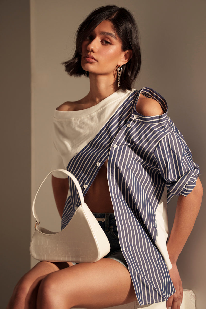 A woman with a short bob hairstyle wears the MIXED UP TSHIRT BLOUSE in blue-and-white stripes with an asymmetrical silhouette, holding a white handbag as she poses seated on a stool against a neutral background.