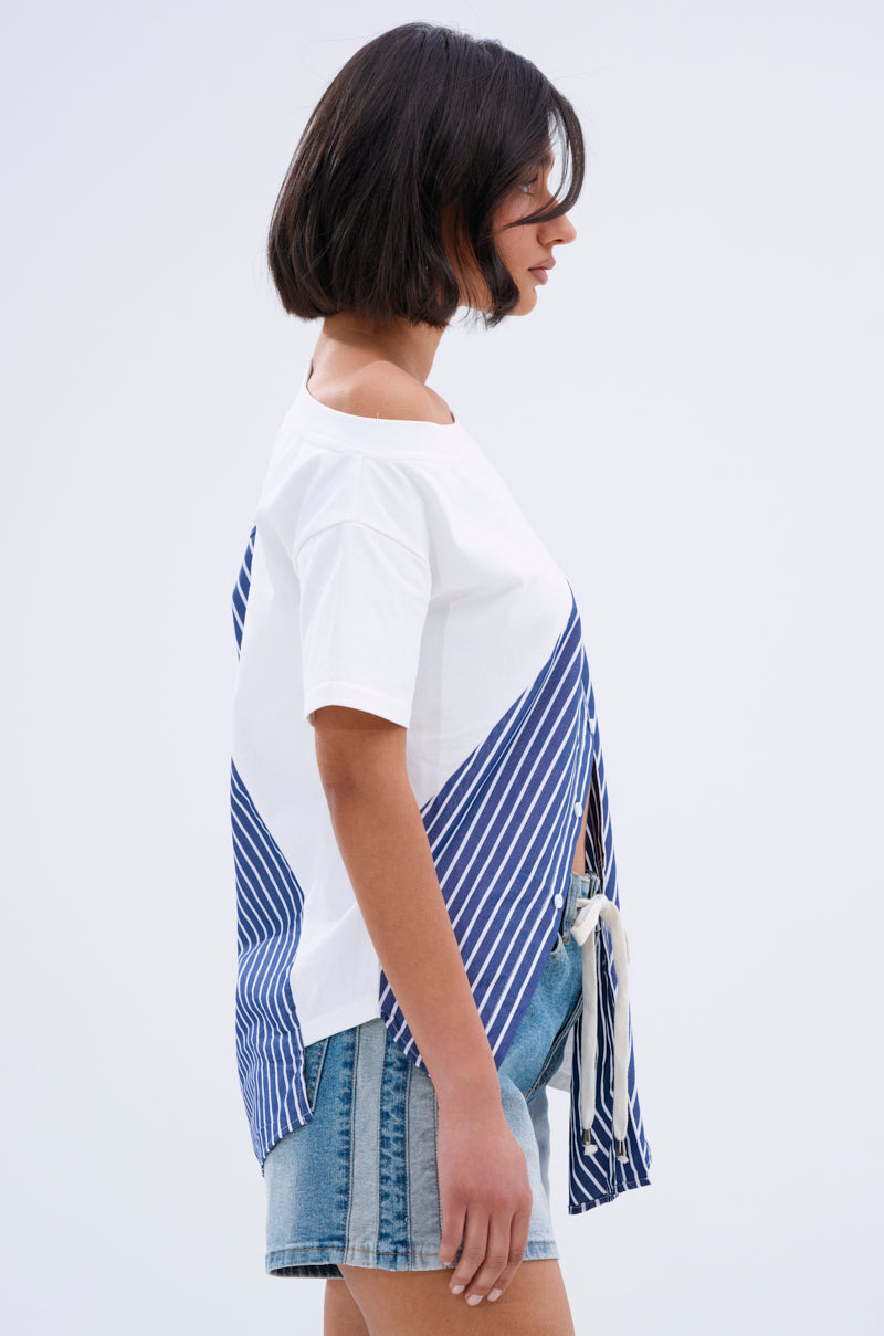 A person with short dark hair stands in profile against a plain light background, wearing the MIXED UP TSHIRT BLOUSE—a white top with blue striped accents—paired with denim shorts.