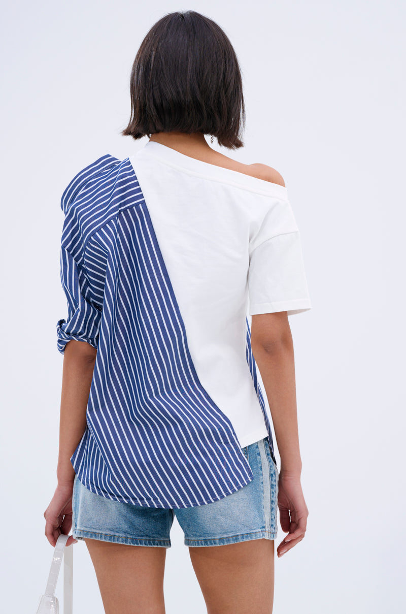 A person with short dark hair, seen from behind, wears the MIXED UP TSHIRT BLOUSE—half blue and white pinstriped, half plain white—with denim shorts and a transparent bag in their left hand.
