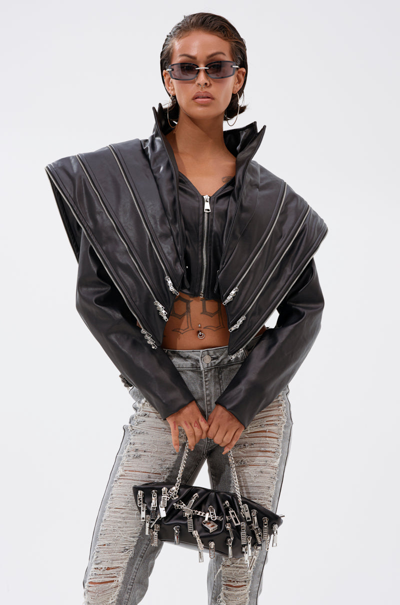 Wearing the COSMO ZIPPER MOTO black faux leather jacket with metallic zippers, ripped jeans, and sunglasses, a person holds a metal charm handbag against a plain white background.