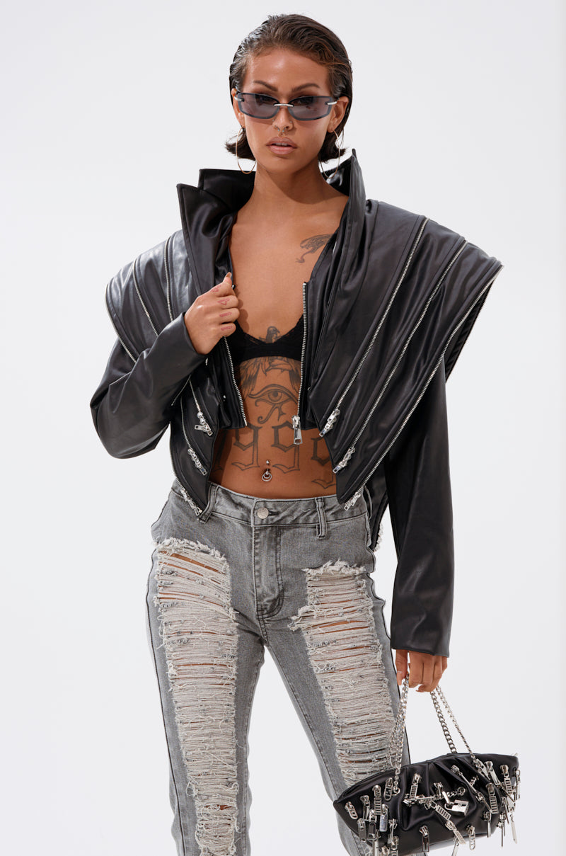 Wearing the COSMO ZIPPER MOTO cropped faux leather jacket over a black bralette and distressed gray jeans, this person holds a small black handbag with silver chains, pairing slicked-back hair and sunglasses for an edgy look.