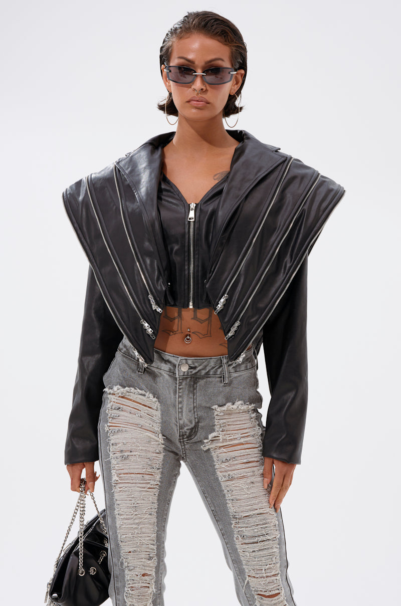 A person wears the COSMO ZIPPER MOTO black cropped jacket with an oversized collar and metallic zippers, gray ripped jeans, sunglasses, and holds a black handbag against a plain white background.