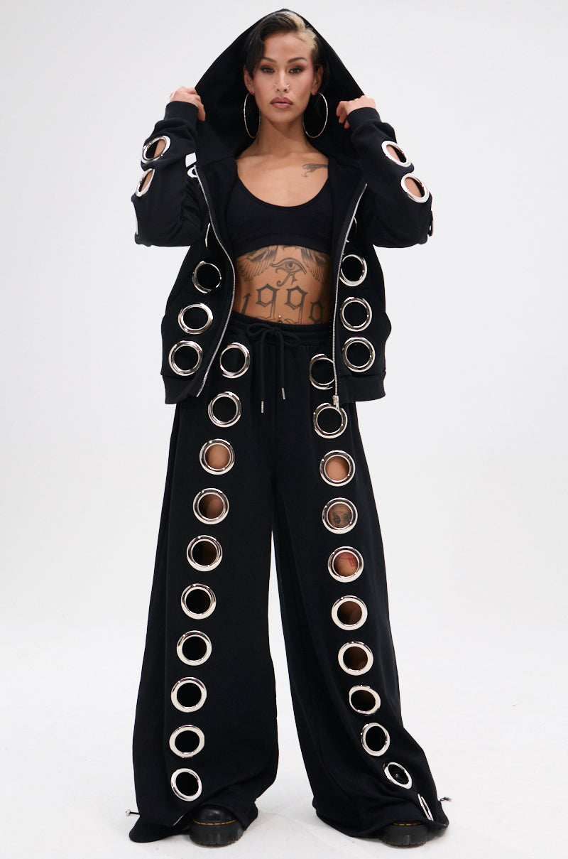 A person poses confidently in THROUGH THE NIGHT WIDE LEG SWEATPANT WITH HOLES, styled with a black hoodie featuring large circular cutouts and silver grommets. They complete the streetwear look with a crop top, hoop earrings, and showcase a tattooed midriff.