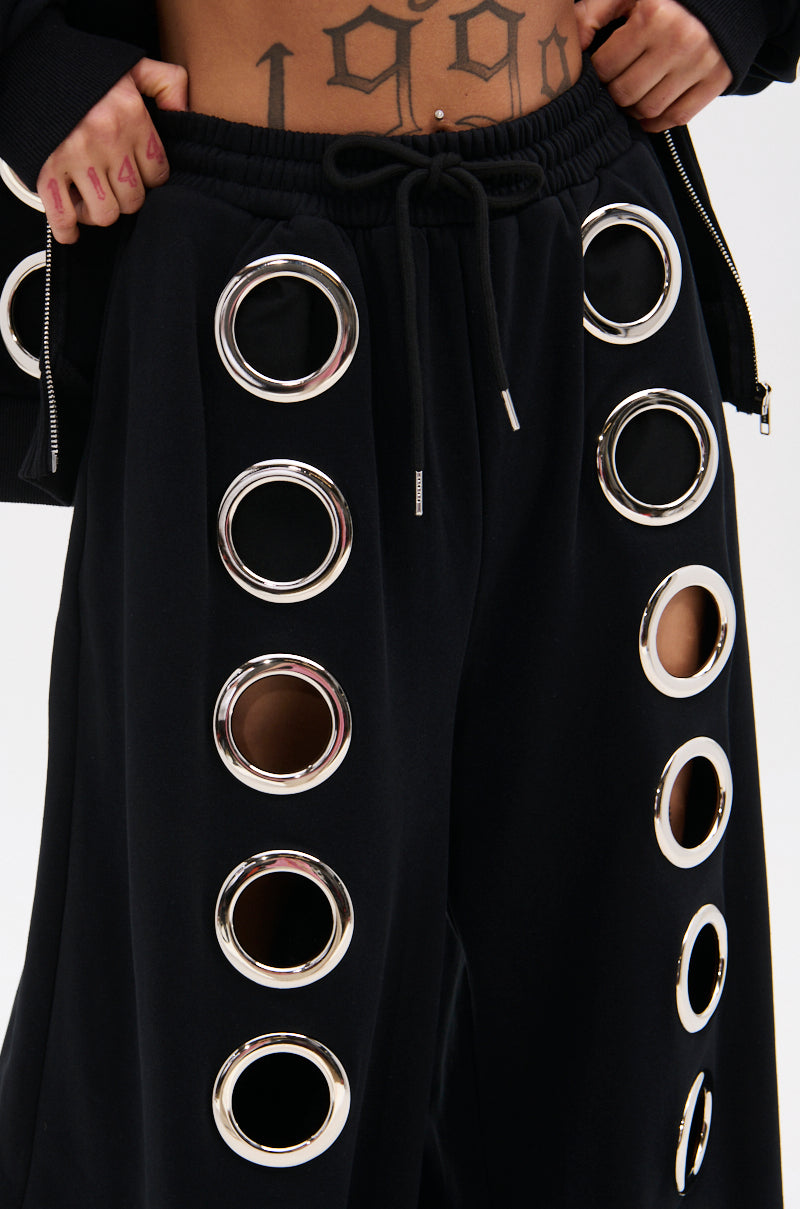 Close-up of a person wearing THROUGH THE NIGHT WIDE LEG SWEATPANT WITH HOLES, featuring black wide legs and large metallic grommet cutouts along the sides. Hands on waistband reveal a tattooed "1990" for a bold streetwear-chic vibe.