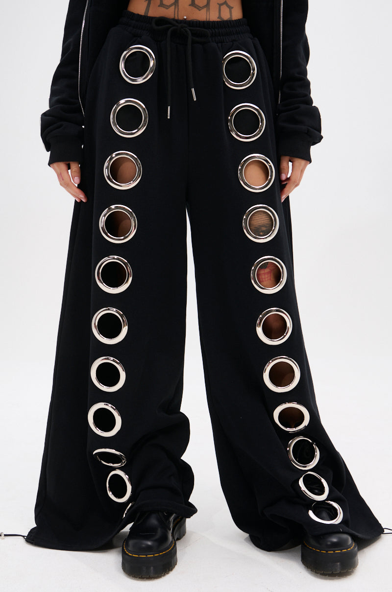 Person wearing THROUGH THE NIGHT WIDE LEG SWEATPANT WITH HOLES—black wide leg sweatpants with silver grommet holes down each leg, styled with black platform shoes for a streetwear-chic look that boldly reveals glimpses of skin.