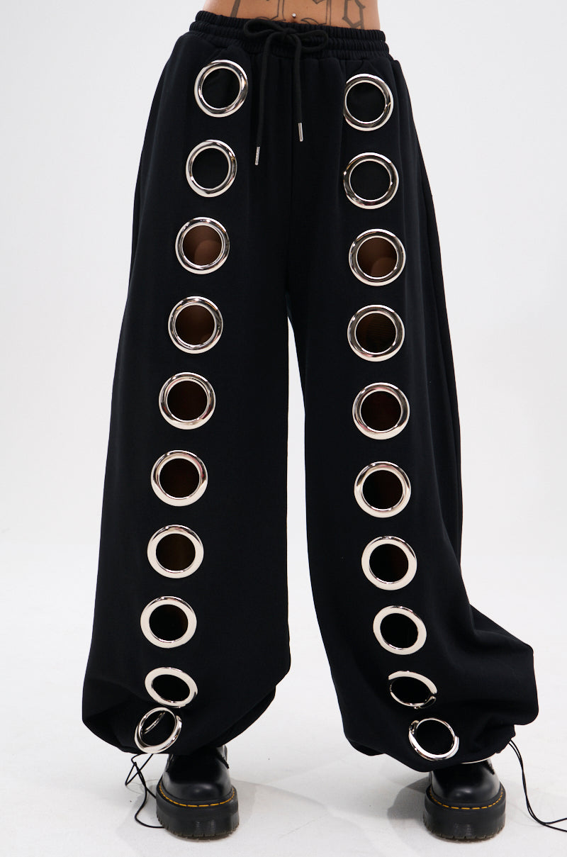 Person wears THROUGH THE NIGHT WIDE LEG SWEATPANT WITH HOLES featuring two vertical rows of large silver grommets down each leg, styled with black shoes for a bold, streetwear-chic look and finished with a drawstring waist.