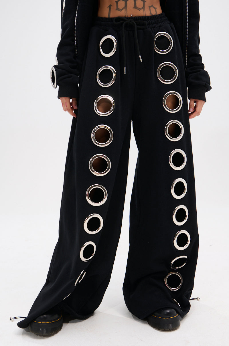 Wearing THROUGH THE NIGHT WIDE LEG SWEATPANT WITH HOLES, featuring large silver grommet cutouts and a drawstring waist, styled with black platform shoes and a sleek black jacket for a streetwear-chic look.