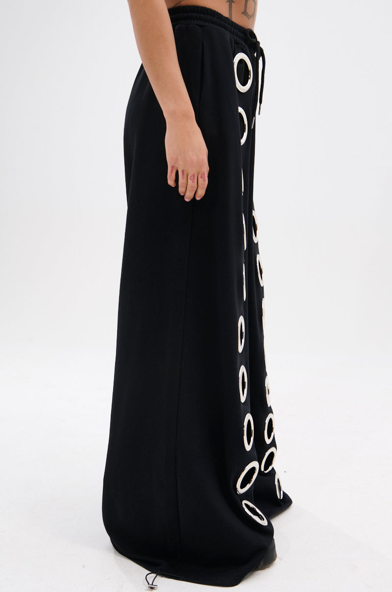 A model wears the THROUGH THE NIGHT WIDE LEG SWEATPANT WITH HOLES, featuring white circular accents down the front, photographed from the side on a plain white background for a streetwear-chic look.