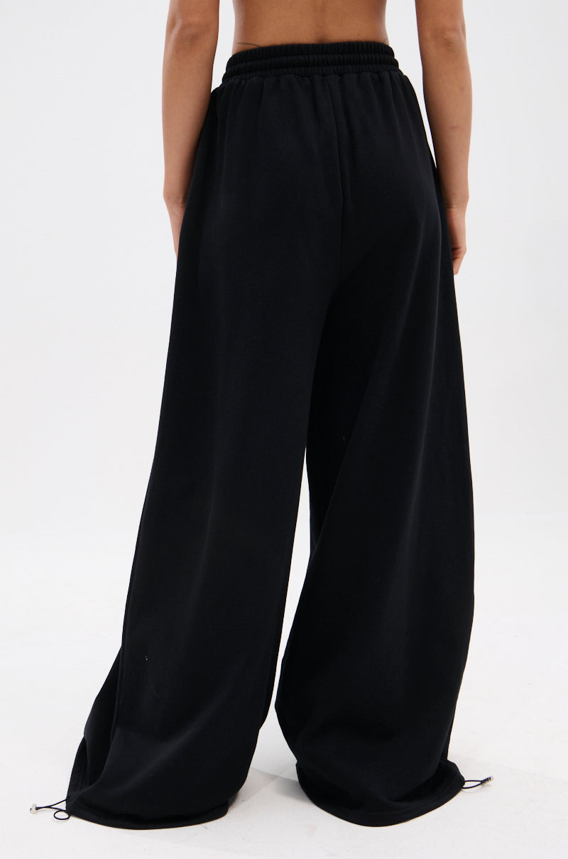 A person is shown from the waist down wearing THROUGH THE NIGHT WIDE LEG SWEATPANT WITH HOLES—loose-fitting with an elastic waistband and ankle drawstrings—showcasing a streetwear-chic vibe against a plain light background.
