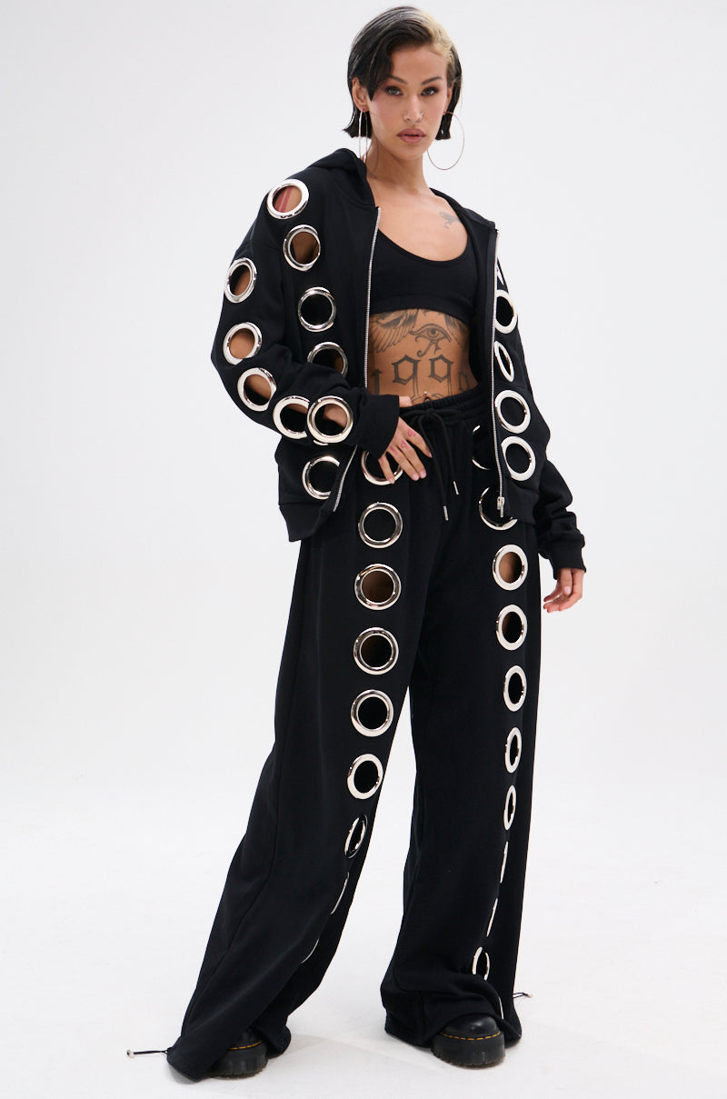 Wearing AKIRA Label’s THROUGH THE NIGHT WIDE LEG SWEATPANT WITH HOLES and a black hoodie with metallic grommets, this person exudes streetwear-chic. Short hair, hoop earrings, and a crop top reveal their tattooed midriff against a white background.