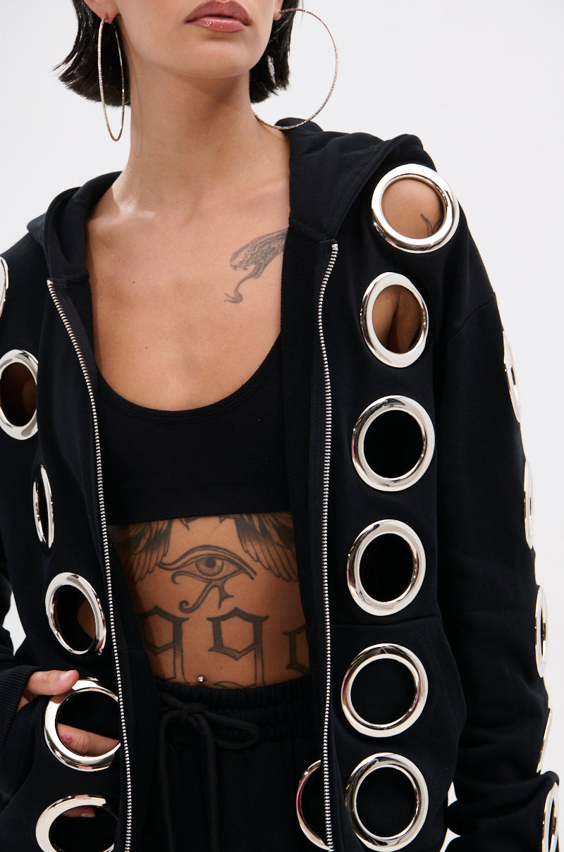 Someone wearing large hoop earrings, a black crop top, and the THROUGH THE NIGHT SWEATSHIRT WITH HOLES, which features oversized metallic grommet holes that reveal a detailed chest tattoo of an eye with wings.