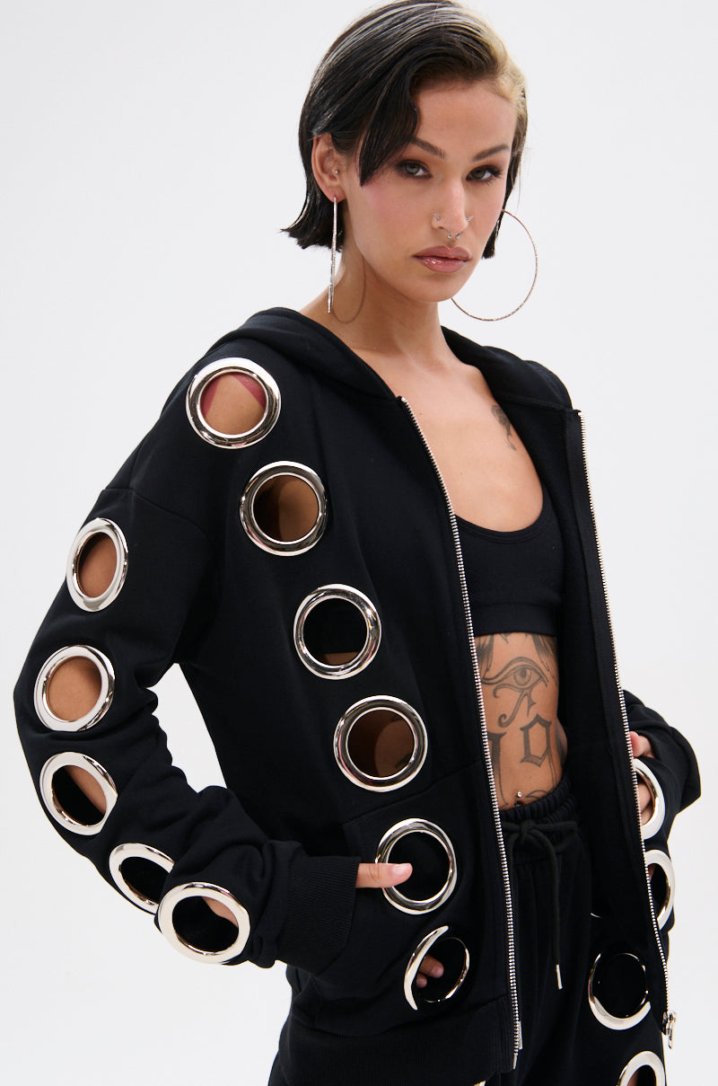 A woman with short, dark hair wears the THROUGH THE NIGHT SWEATSHIRT WITH HOLES, a zip-up hoodie featuring large metallic grommet holes on the sleeves and front. She pairs it with a black crop top, black pants, hoop earrings, and reveals a torso tattoo.