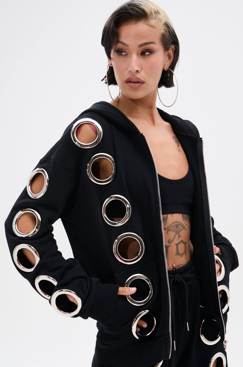 A woman wears the THROUGH THE NIGHT SWEATSHIRT WITH HOLES—a black zip-up hoodie with large metallic grommet holes on the sleeves—styled with a black sports bra, black pants, hoop earrings, visible tattoos, and short hair featuring a blonde streak.