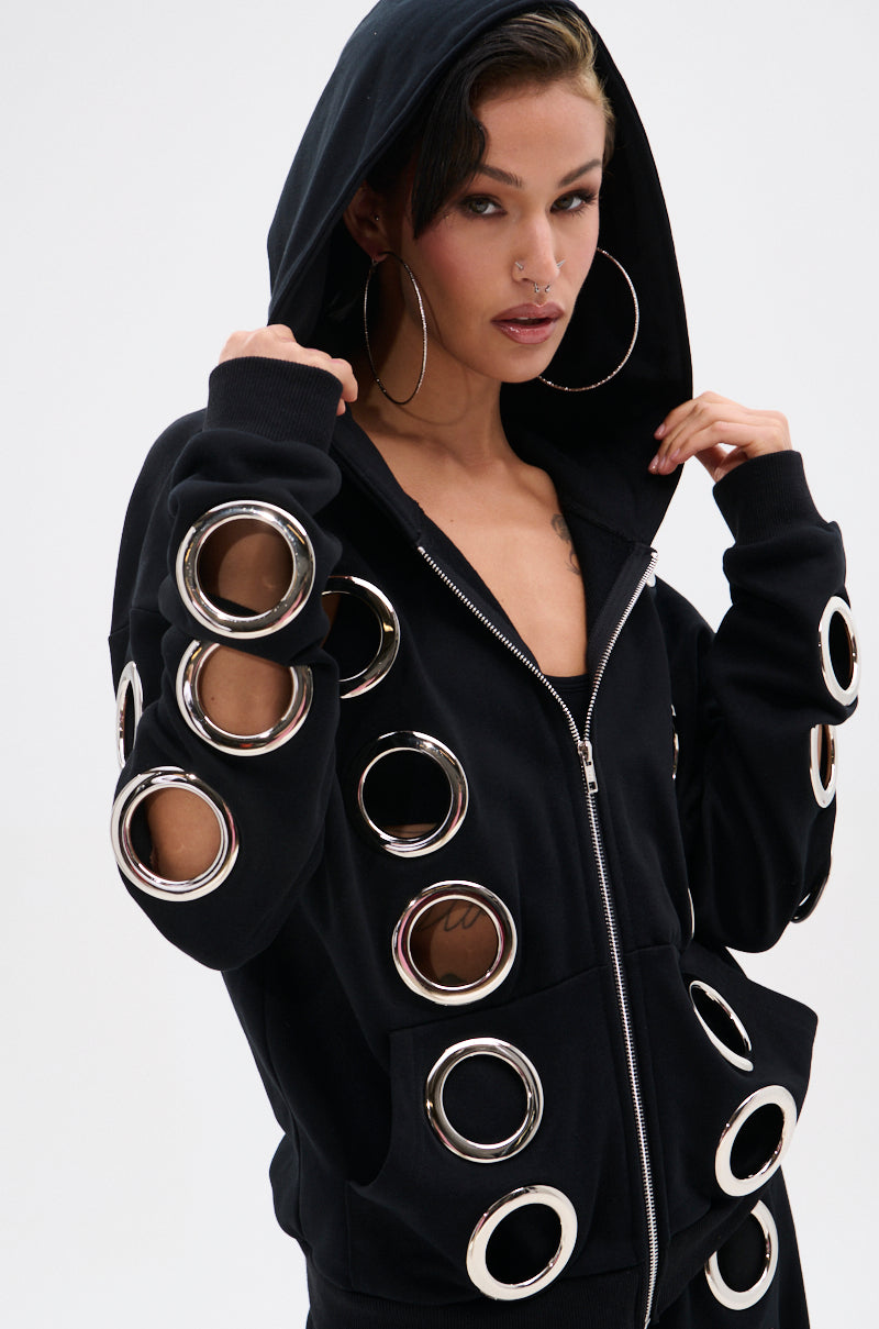 A woman models the THROUGH THE NIGHT SWEATSHIRT WITH HOLES zip-up hoodie, featuring large metallic grommet holes, hood up and hoop earrings, posing confidently against a plain white background.
