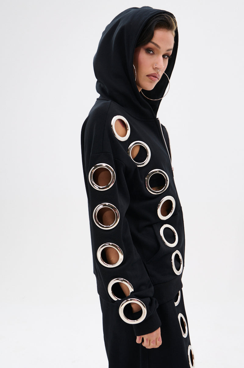 A woman in the THROUGH THE NIGHT SWEATSHIRT WITH HOLES zip-up hoodie, featuring oversized metal grommet holes along the sleeves and sides, stands against a plain white background with her hood up, looking at the camera.