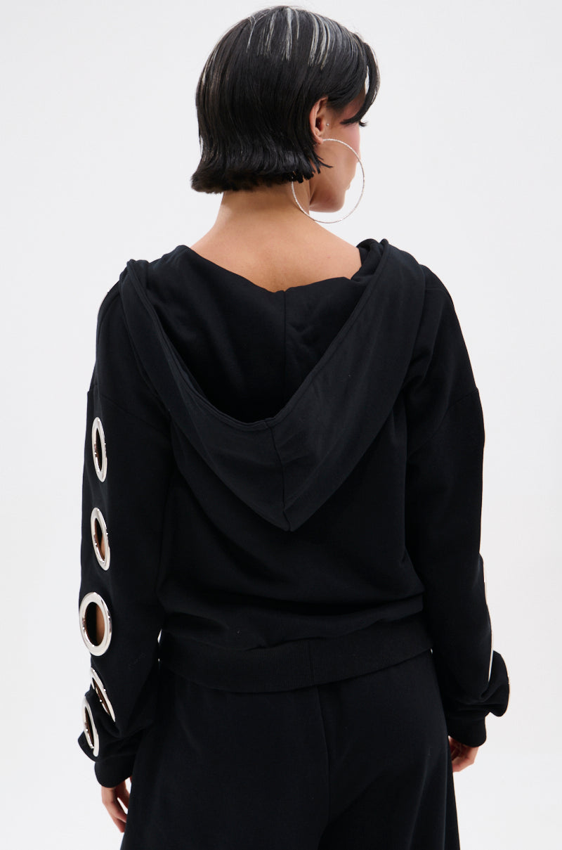 A person with short dark hair and hoop earrings is seen from the back wearing the THROUGH THE NIGHT SWEATSHIRT WITH HOLES zip-up hoodie, featuring large metallic grommet holes on the sleeves, paired with matching black pants.