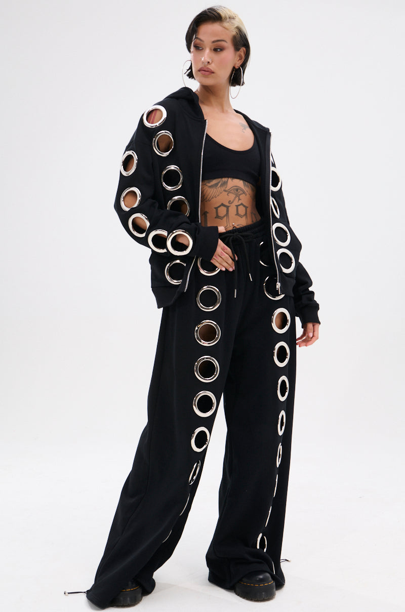 A person wears the THROUGH THE NIGHT SWEATSHIRT WITH HOLES—black with metallic grommet holes and large circular cutouts—paired with black pants, a crop top, and shoes, standing against a plain white background.