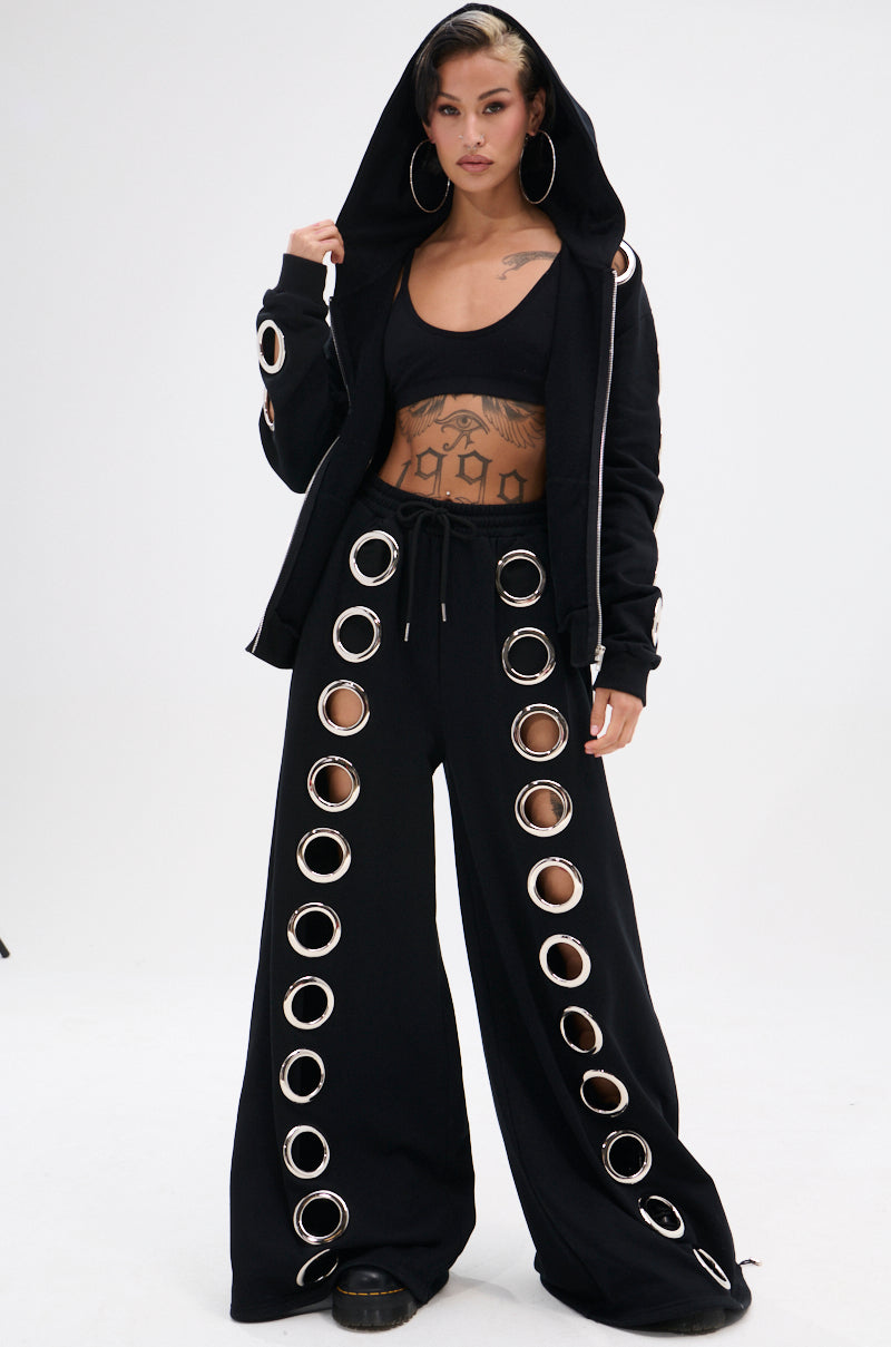 Someone stands against a plain white background wearing the THROUGH THE NIGHT SWEATSHIRT WITH HOLES, a black zip-up hoodie featuring large metallic grommet holes, paired with wide-leg pants and a black crop top.
