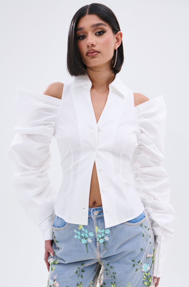A woman with straight, shoulder-length dark hair wears the CRAZY IN LOVE BUTTON DOWN BLOUSE, a white cold-shoulder top with statement sleeves, paired with light blue floral-embroidered jeans against a plain light background.