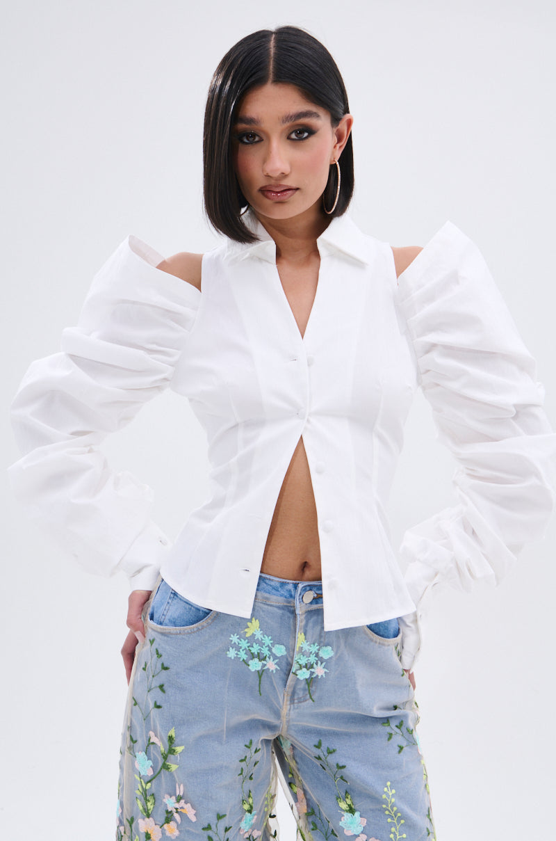 A woman with straight dark hair wears the CRAZY IN LOVE BUTTON DOWN BLOUSE, featuring statement sleeves and cold shoulder cutouts, paired with light blue jeans with colorful floral embroidery. She poses confidently against a plain background.