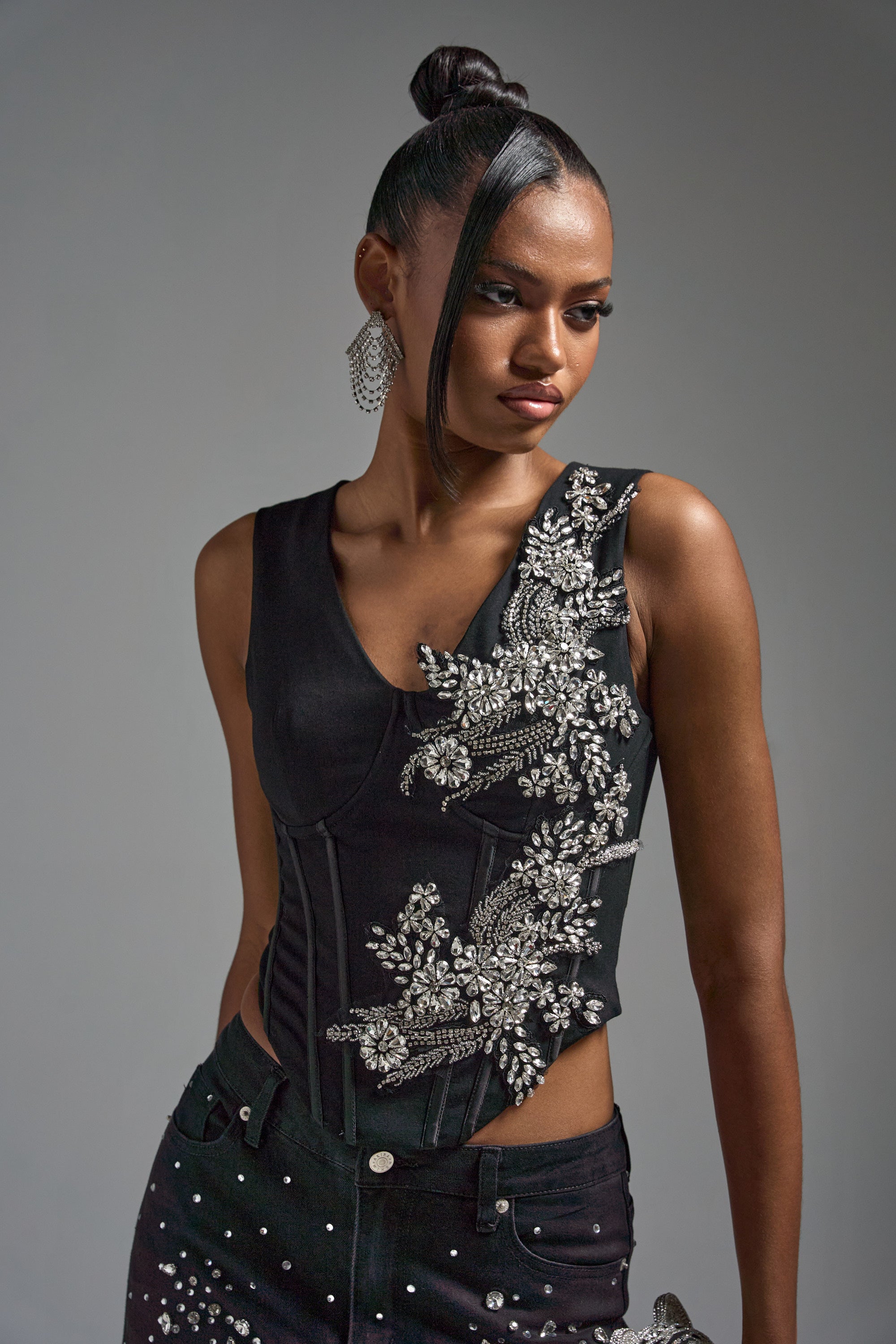 A woman with a high bun models the LOTS OF LOVE TO GIVE RHINESTONE BLOUSE, a black cropped top detailed with silver rhinestones, paired with matching embellished pants. She looks to the side against a plain gray background.