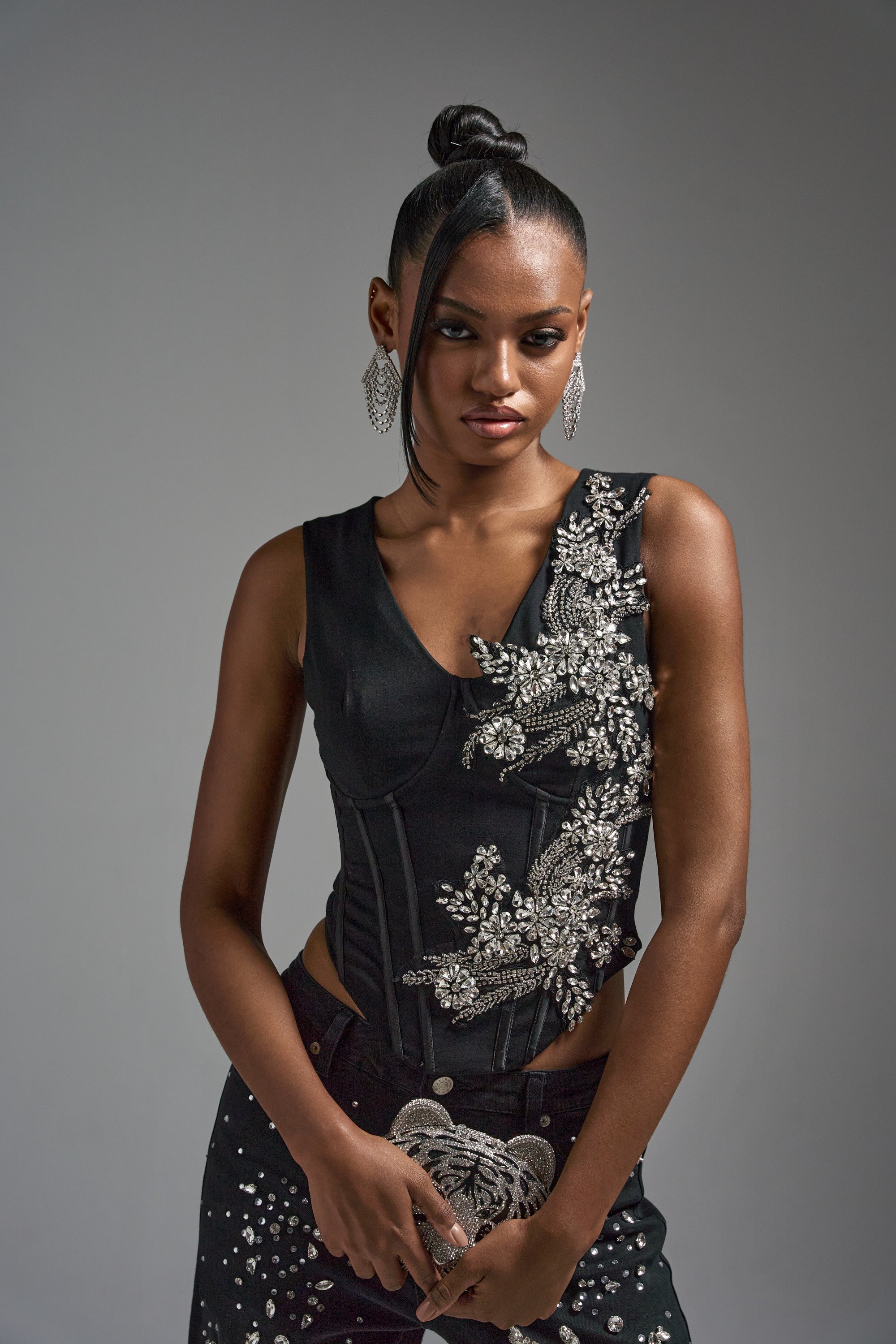 A woman stands against a gray background, wearing the LOTS OF LOVE TO GIVE RHINESTONE BLOUSE adorned with silver beadwork, matching earrings, and holding a sparkling clutch. Her hair is styled in a high bun with loose bangs.