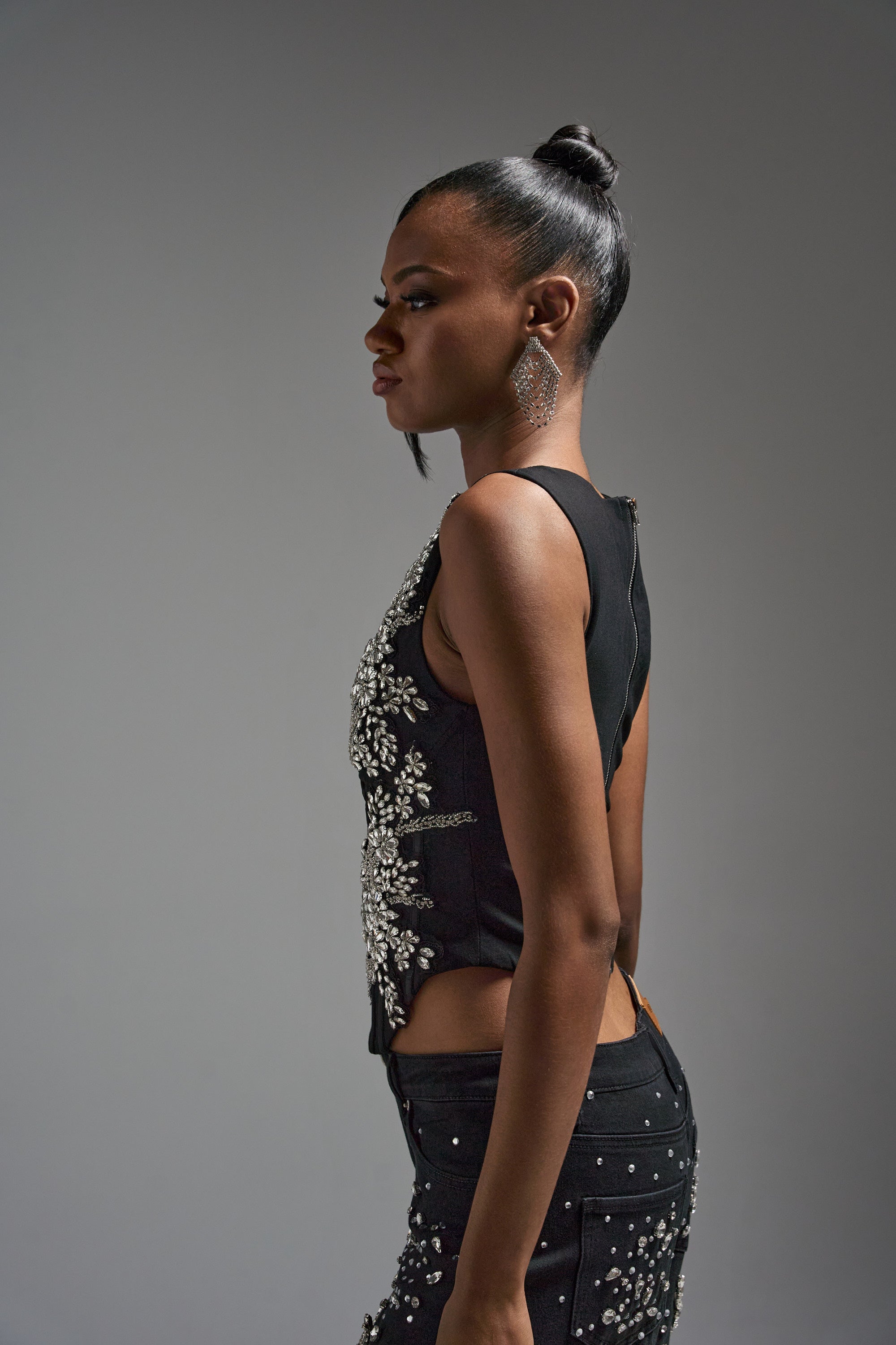 A woman with dark skin and straight hair in a bun stands in profile against a plain background, wearing the LOTS OF LOVE TO GIVE RHINESTONE BLOUSE, paired with pants featuring silver embellishments and large geometric earrings.