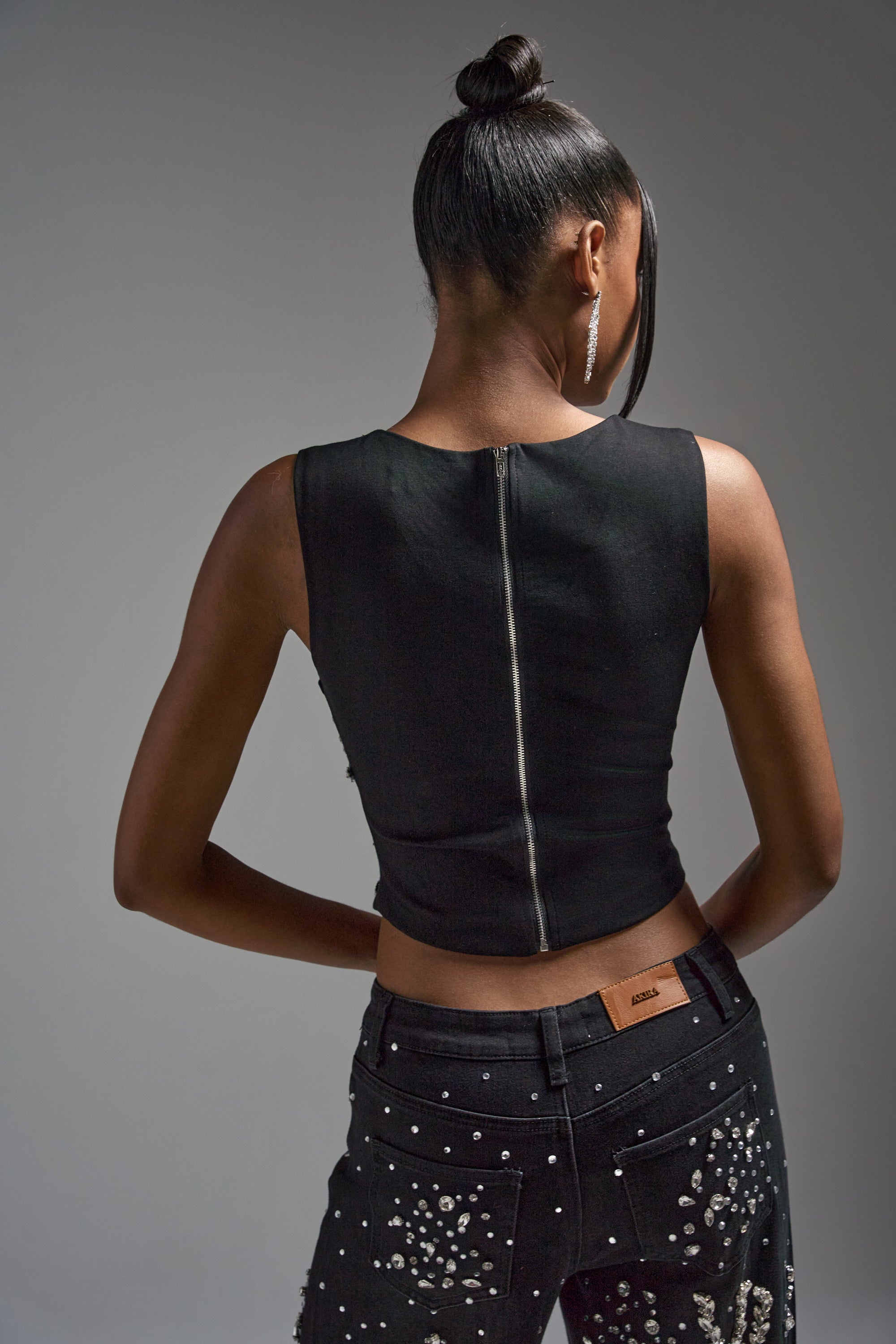 A woman stands with hands on hips, wearing the LOTS OF LOVE TO GIVE RHINESTONE BLOUSE and black jeans adorned with pearls and rhinestones. Her hair is styled in a neat bun, and the background is gray.