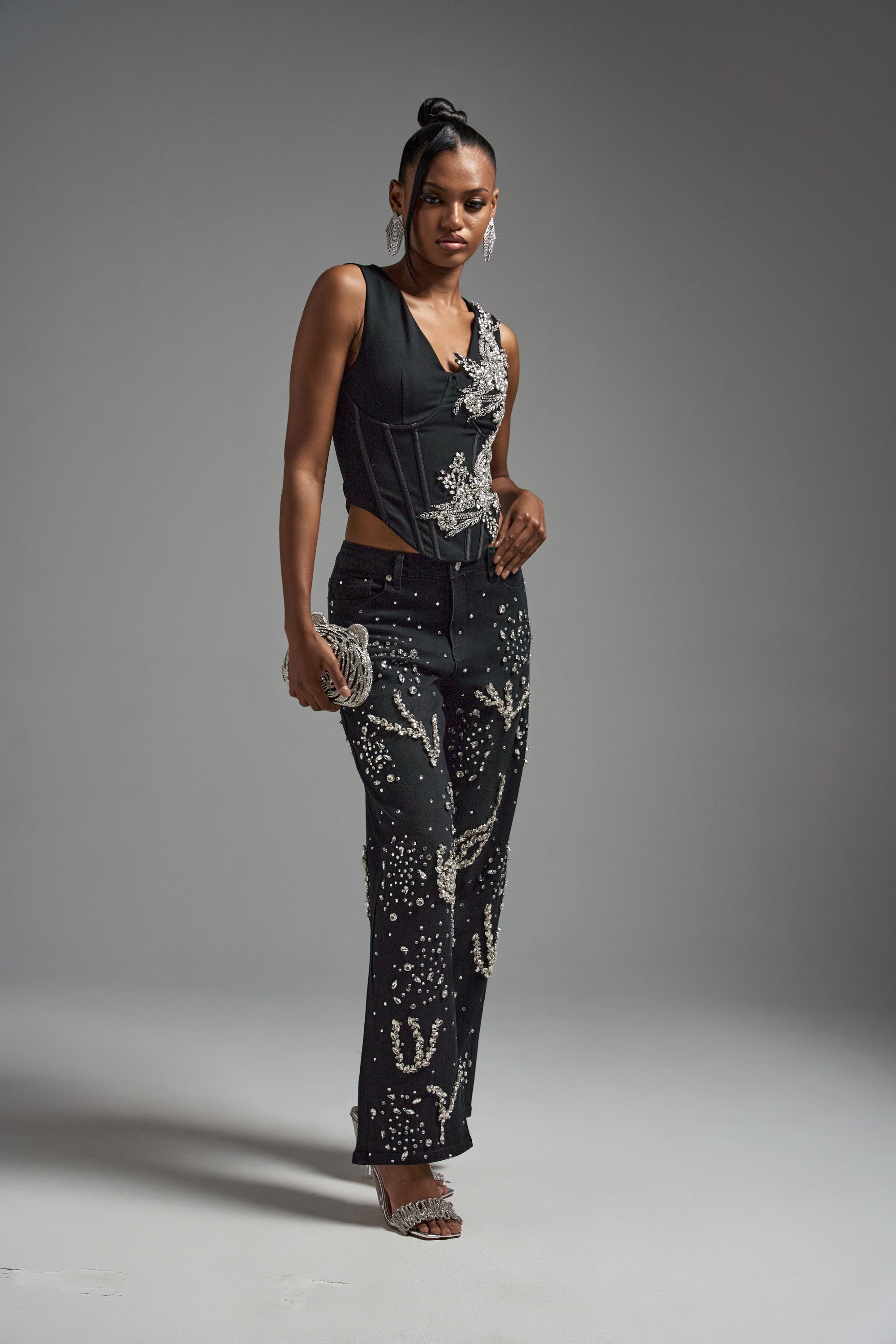 A woman models the LOTS OF LOVE TO GIVE RHINESTONE BLOUSE with high-waisted black pants featuring silver accents. She pairs the look with silver heels, large earrings, and a silver clutch, set against a plain gray background.