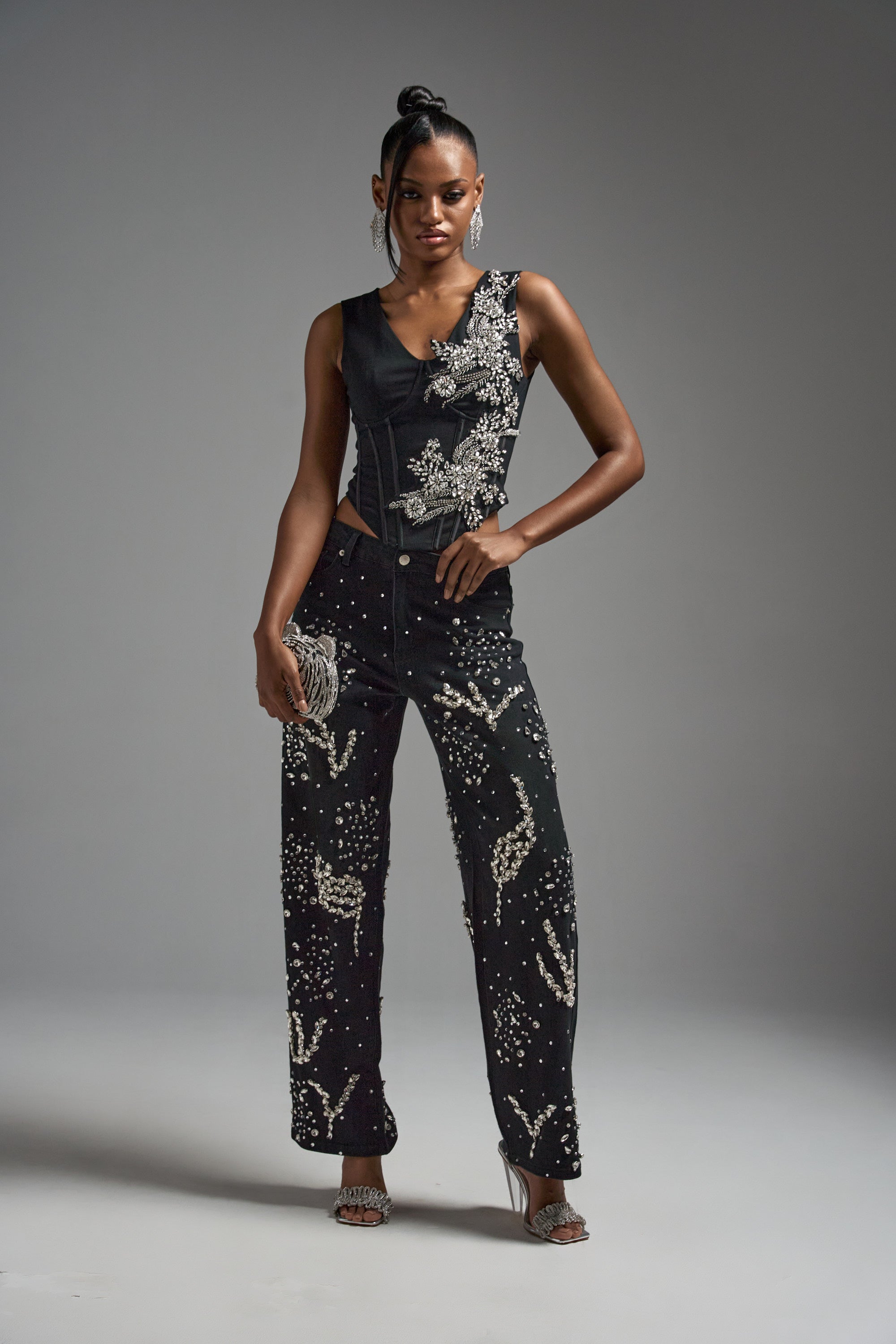 A woman stands confidently in the LOTS OF LOVE TO GIVE RHINESTONE BLOUSE, a sleeveless black top adorned with white beaded patterns, paired with matching black pants, heels, statement earrings, and a clutch, set against a plain gray background.