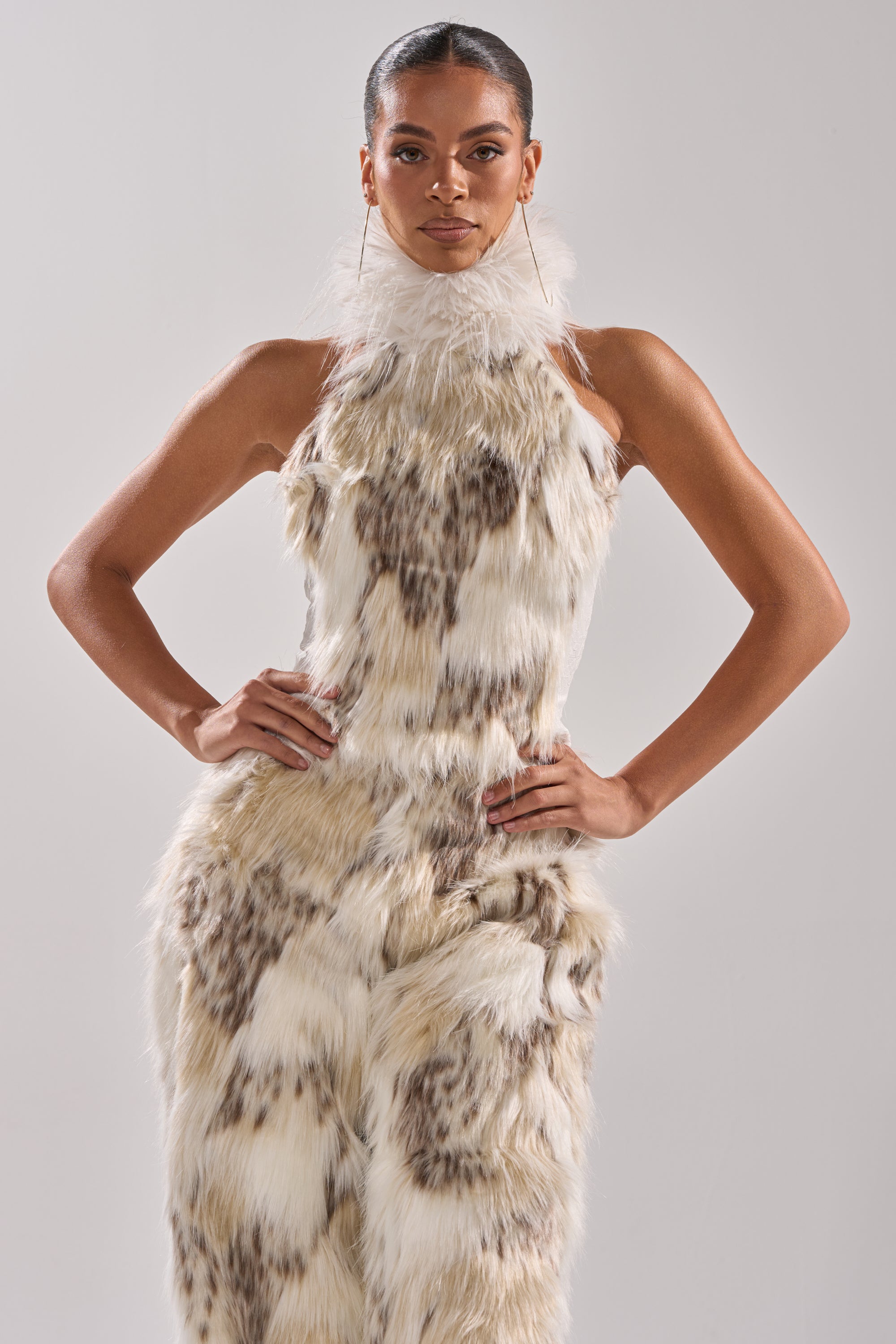 A person stands confidently with hands on hips in a sleeveless, high-neck white and brown faux fur FLINTSTONES MOCK NECK FUR TOP against a plain light gray background.