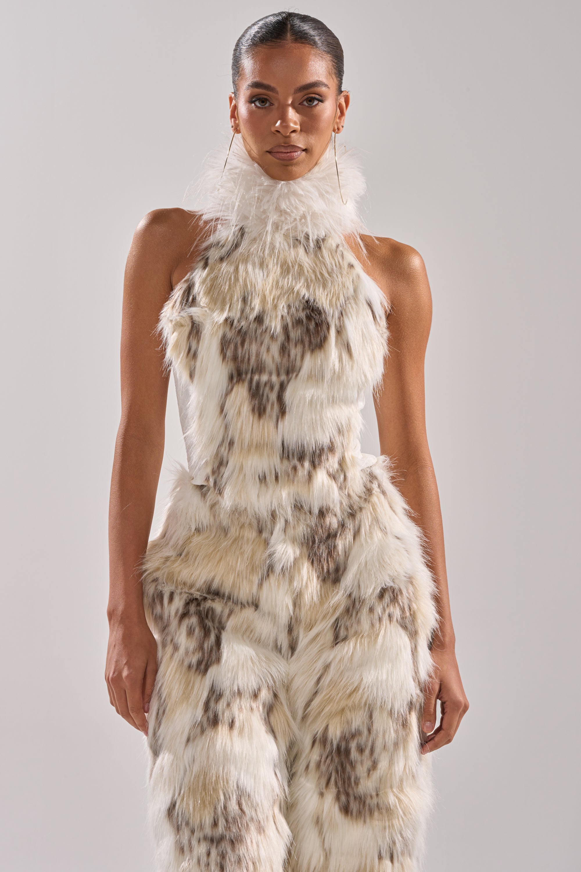 A woman models the dramatic, sleeveless FLINTSTONES MOCK NECK FUR TOP with a high neck and fluffy faux fur texture, standing against a plain light gray background for a unique fashion statement.
