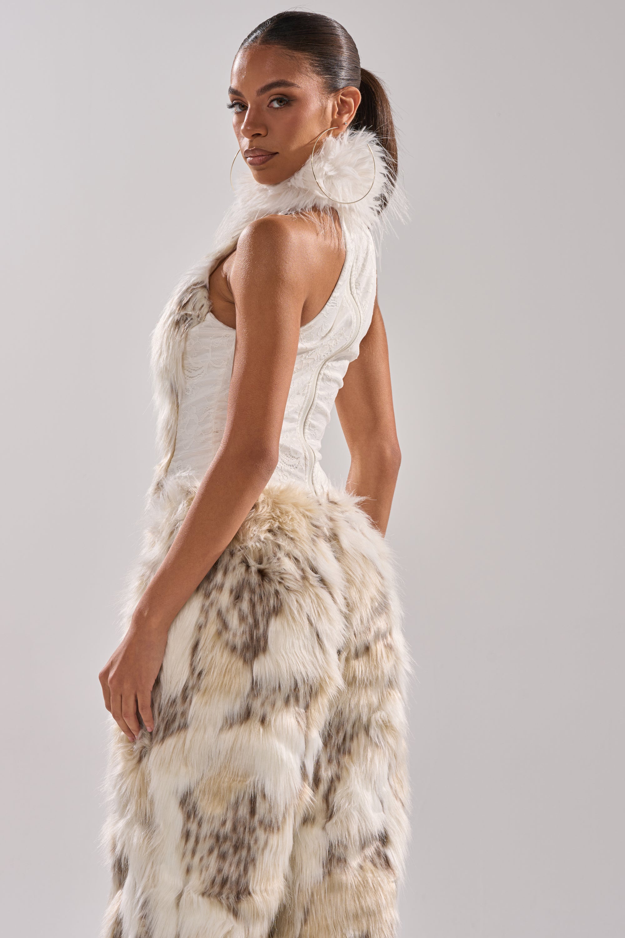 A woman with a sleek ponytail wears the sleeveless white AKIRA Label FLINTSTONES MOCK NECK FUR TOP, posing sideways and gazing at the camera against a plain, light gray background.