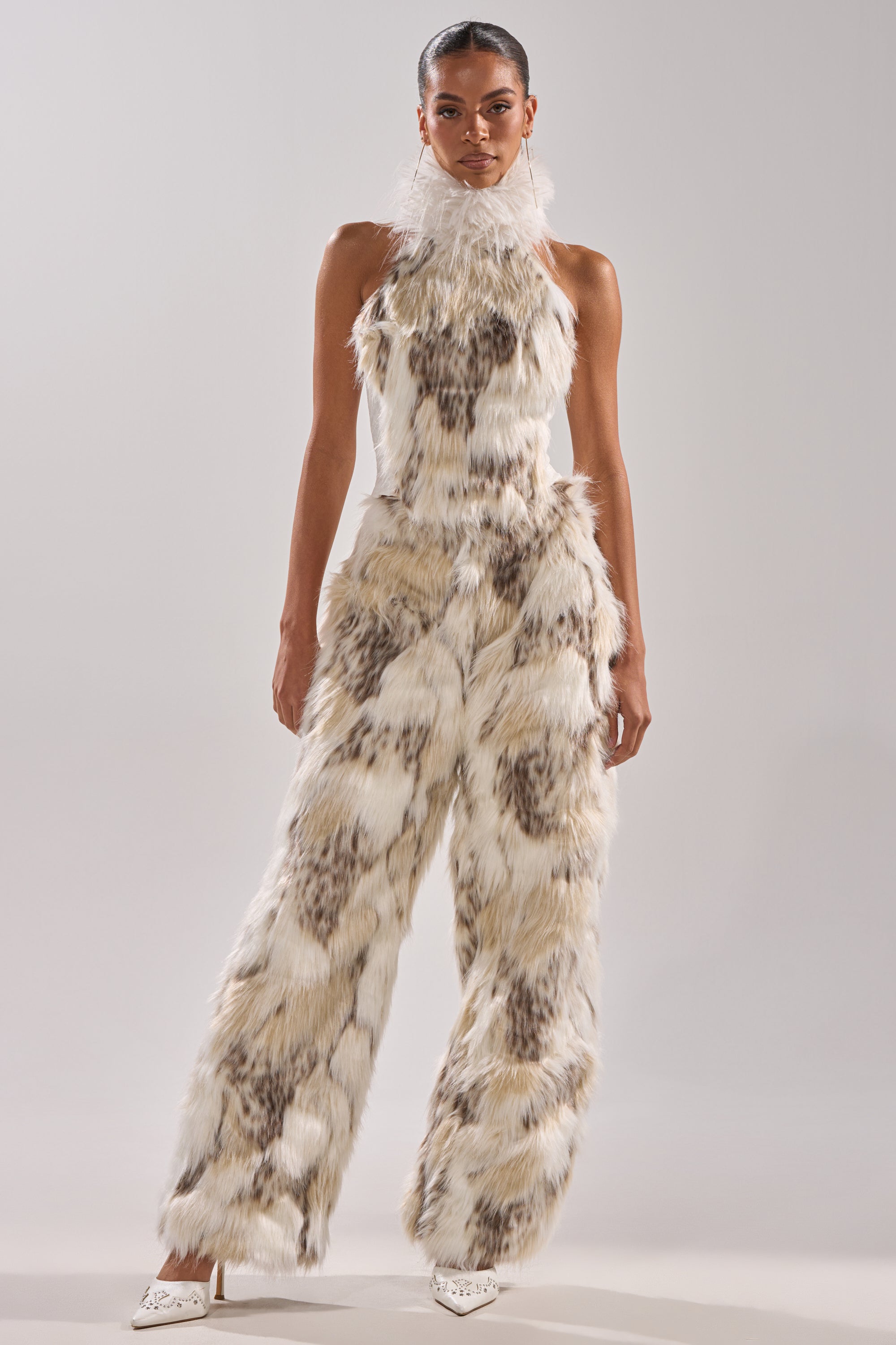 A woman poses confidently in the FLINTSTONES MOCK NECK FUR TOP, a sleeveless piece with a spotted cream, brown, and gray faux fur texture and sheer ruffled collar, styled with white pointed-toe heels against a light gray background.
