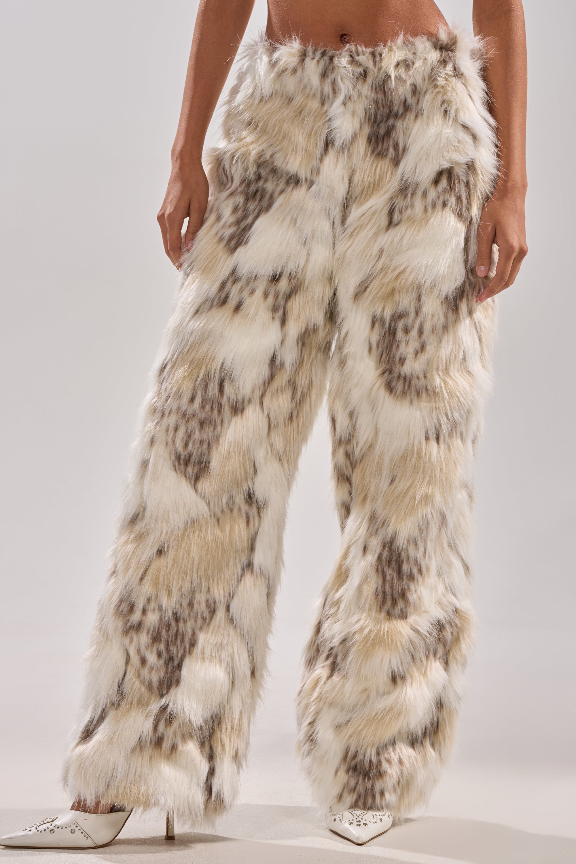 A person stands in FLINTSTONES FUR PANTS—high-waisted, wide-leg faux fur with a spotted cream, brown, and white pattern—paired with white pointed-toe heels. The photo is cropped at the shoulders.