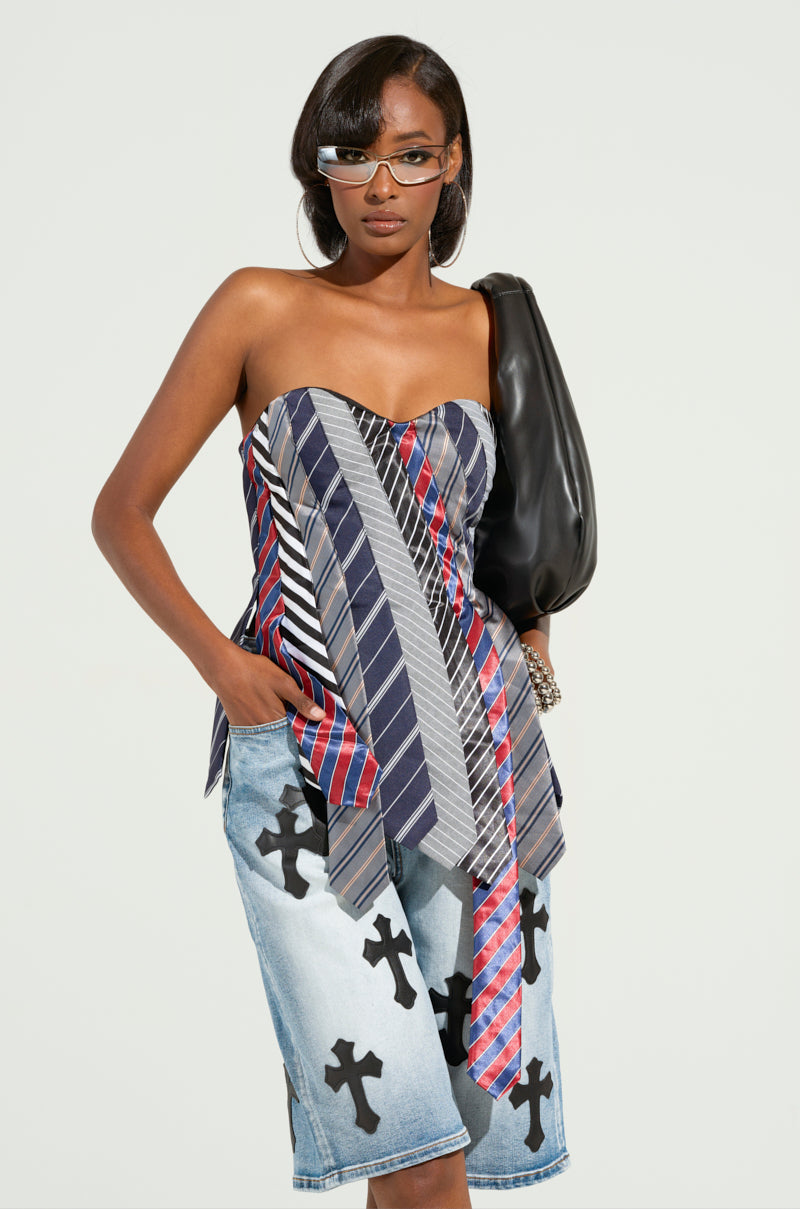 A person wears the STRICTLY BUSINESS TIE DETAIL CORSET TOP, a strapless piece with a distinctive design, paired with knee-length denim shorts adorned with cross motifs. They accessorize with a black bag and stylish sunglasses.