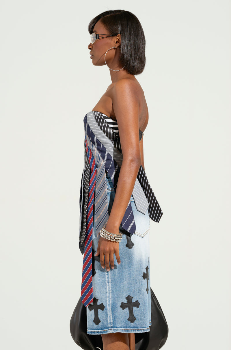 A side view of a person wearing the STRICTLY BUSINESS TIE DETAIL CORSET TOP, crafted from various neckties with a sweetheart neckline, paired with a denim skirt featuring cross designs. The individual has short hair, hoop earrings, and sunglasses, accessorized with bracelets against a plain backdrop.