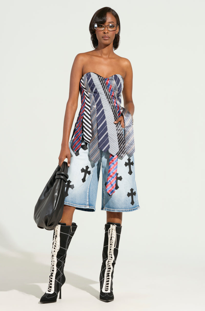 A person wearing the STRICTLY BUSINESS TIE DETAIL CORSET TOP, denim shorts with cross patterns, and knee-high boots. They are carrying a large black bag and wearing sunglasses, all set against a light-colored background.