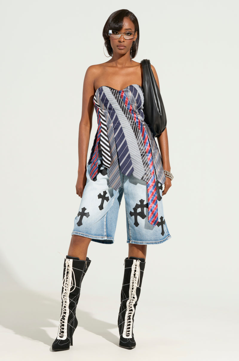 A woman stands confidently against a plain background, wearing the STRICTLY BUSINESS TIE DETAIL CORSET TOP with a patchwork design made from various neckties, paired with denim shorts featuring cross patterns, lace-up knee-high boots, visor sunglasses, and carrying a black shoulder bag.