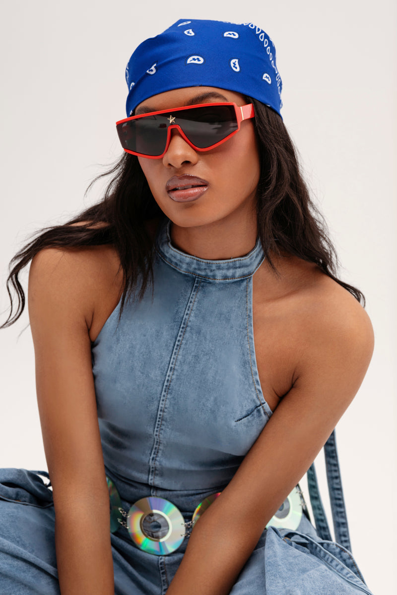 A person is dressed in a blue denim halter top and skirt, a paisley-patterned blue bandana, and wears RED HOT SUNGLASSES with dark-tinted lenses. The outfit is completed with a belt adorned with shiny circular discs.