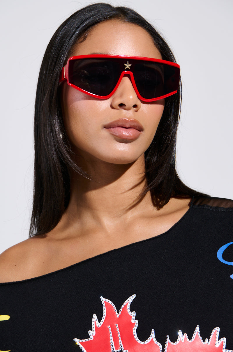 A person with long dark hair wears RED HOT SUNGLASSES, which are oversized and feature a star embellishment. They don a black shirt with colorful patterns, and their distinctive monobrow silhouette pops against the plain white background.