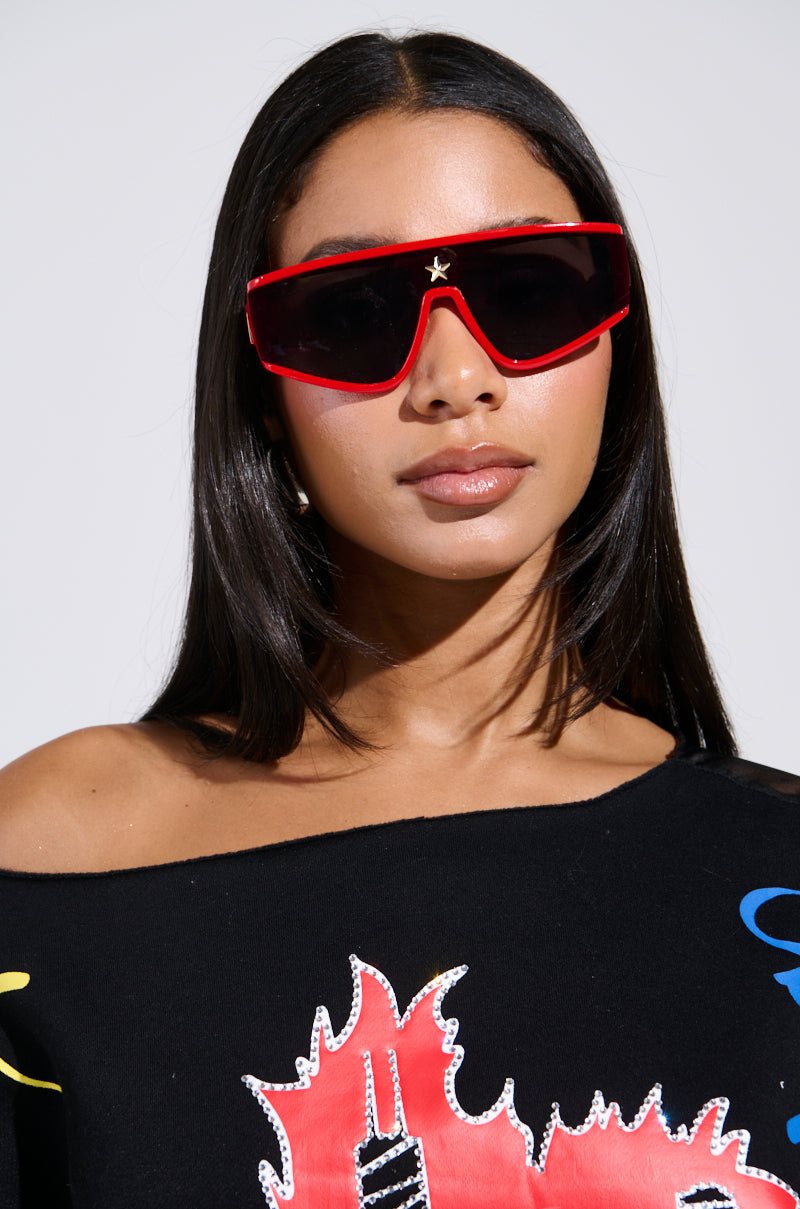 A woman with long dark hair wears RED HOT SUNGLASSES and an off-shoulder black top with a graphic design, gazing forward against a plain background.