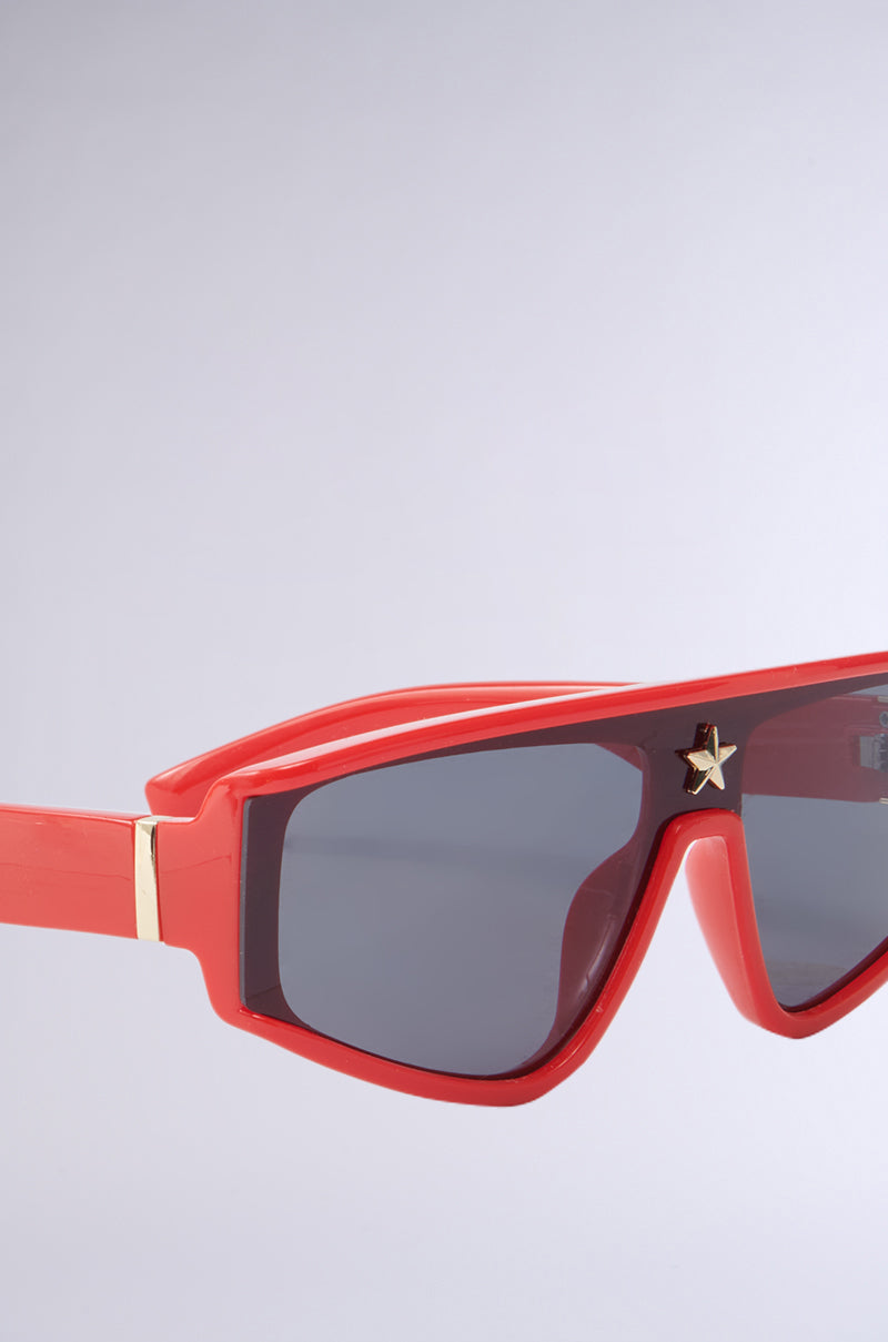A close-up of RED HOT SUNGLASSES with angular frames and dark-tinted lenses; a small gold star decorates one lens. The background is light gray.