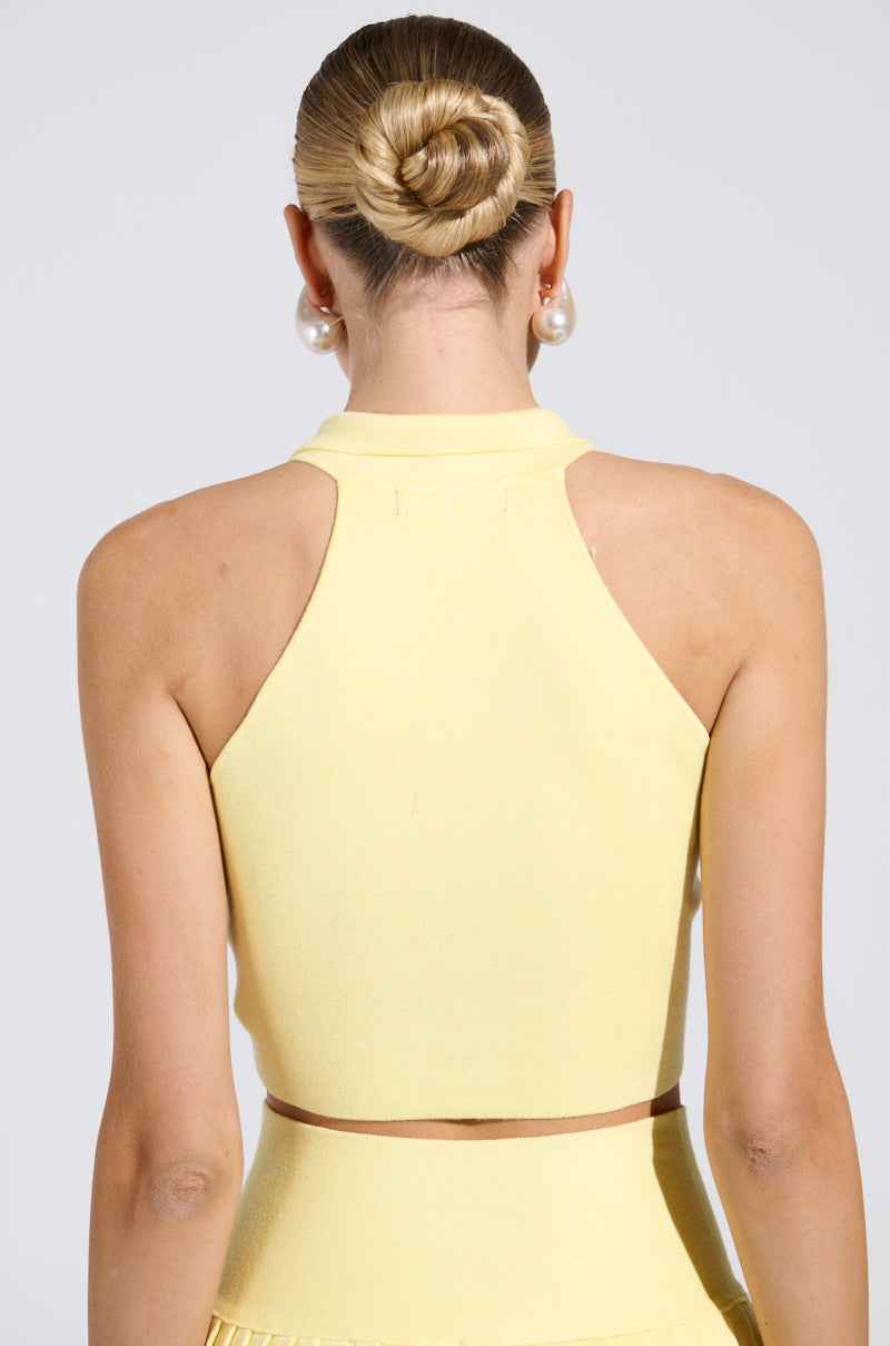 Rear view of a person with blonde hair in a sleek bun, wearing the Lemony Emily Cropped Sweater Vest in yellow. This ensemble features a sleeveless silhouette with large pearl earrings and is paired with a matching skirt, standing out against the plain light-colored background.
