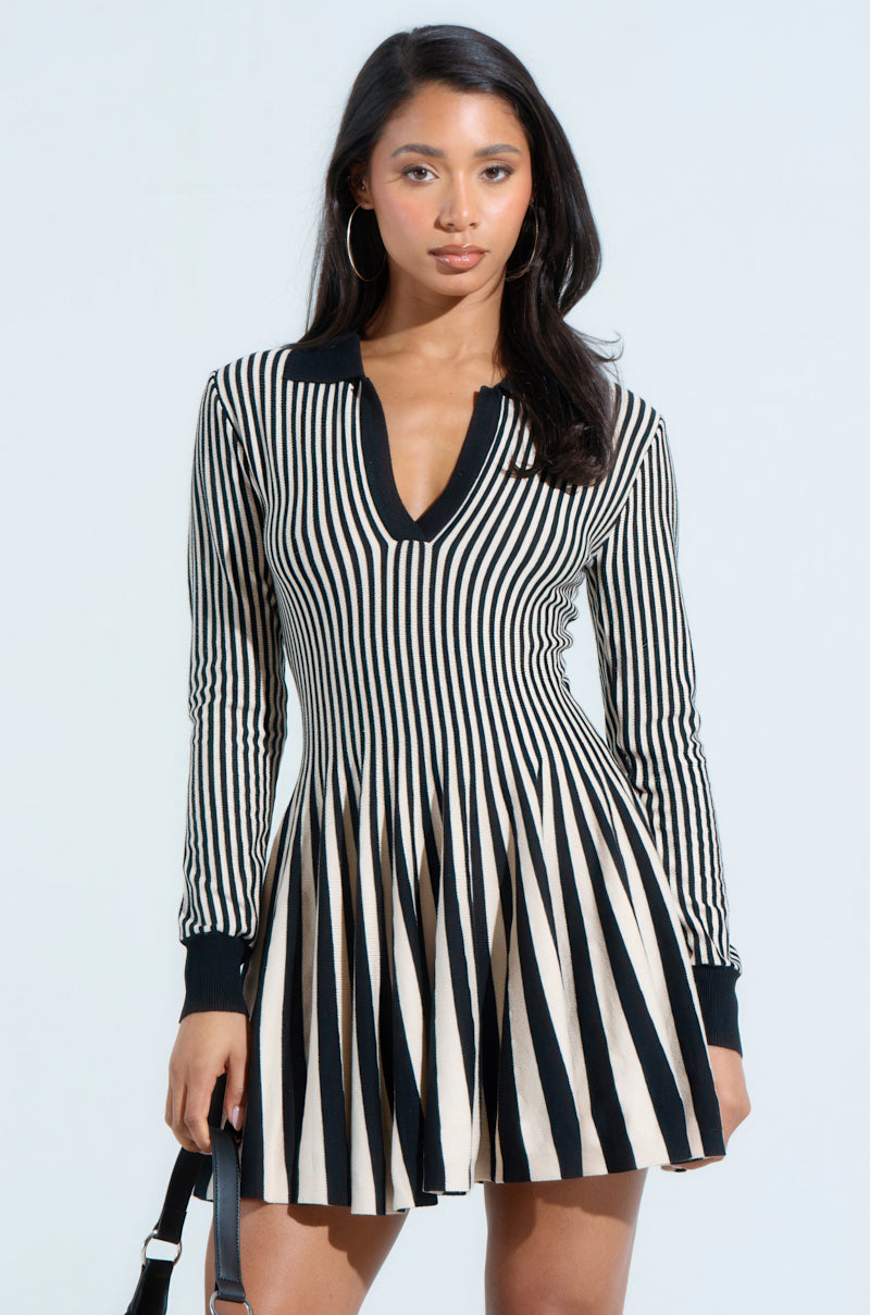 A woman poses gracefully in an ALINA pleated knit fit and flare mini dress with long sleeves, featuring a black and white color block design, accessorized with a chic black bag and hoop earrings. Her long hair cascades elegantly against a plain backdrop.