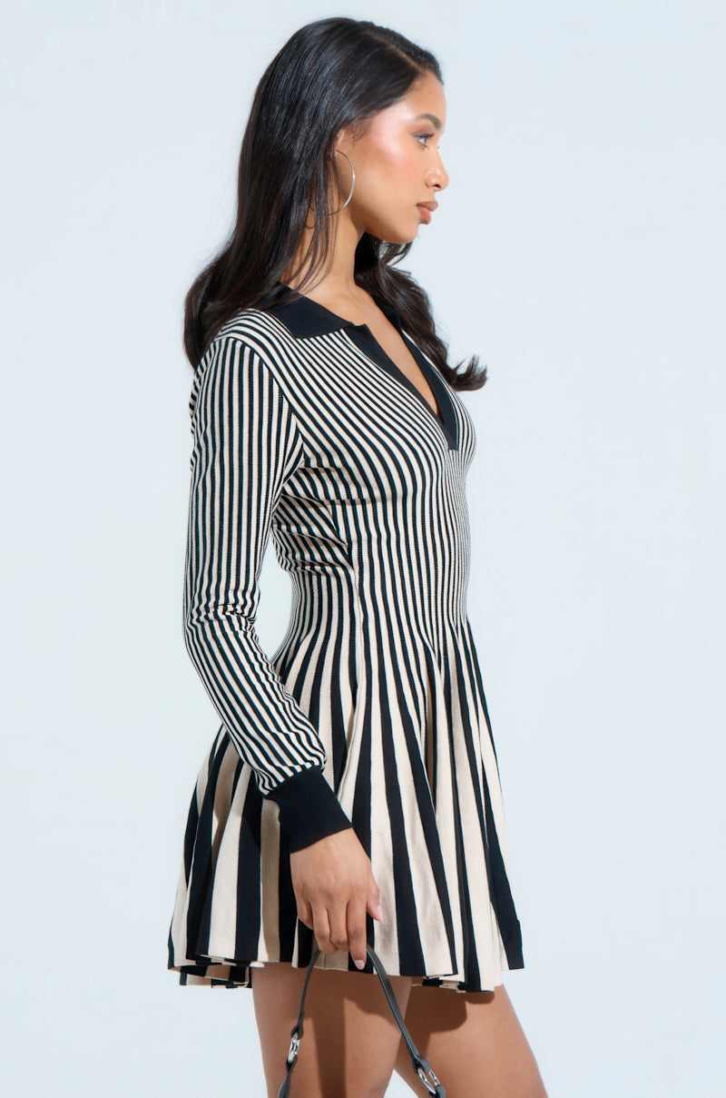 A woman with long dark hair is wearing the ALINA Pleated Knit Fit and Flare Mini Dress with long sleeves, showcasing a stylish color block pattern. She holds a small bag in her left hand and stands gracefully turned against a plain background.