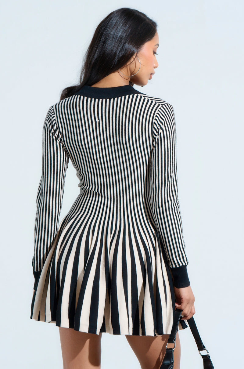 A woman with long dark hair faces away, donning the ALINA pleated knit fit and flare mini dress in black and white stripes. She holds a black handbag and wears hoop earrings against a plain light background.