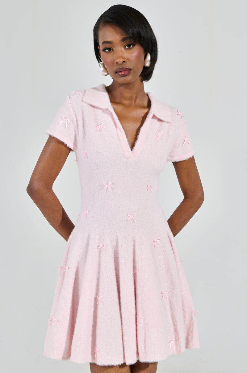A person in the BOW DOWN GIRL MINI DRESS IN PINK, featuring a flared skirt and small bow details, poses with hands behind their back. They have short dark hair and large pearl earrings, and the dress has a light pink color with a V-neck and collar.